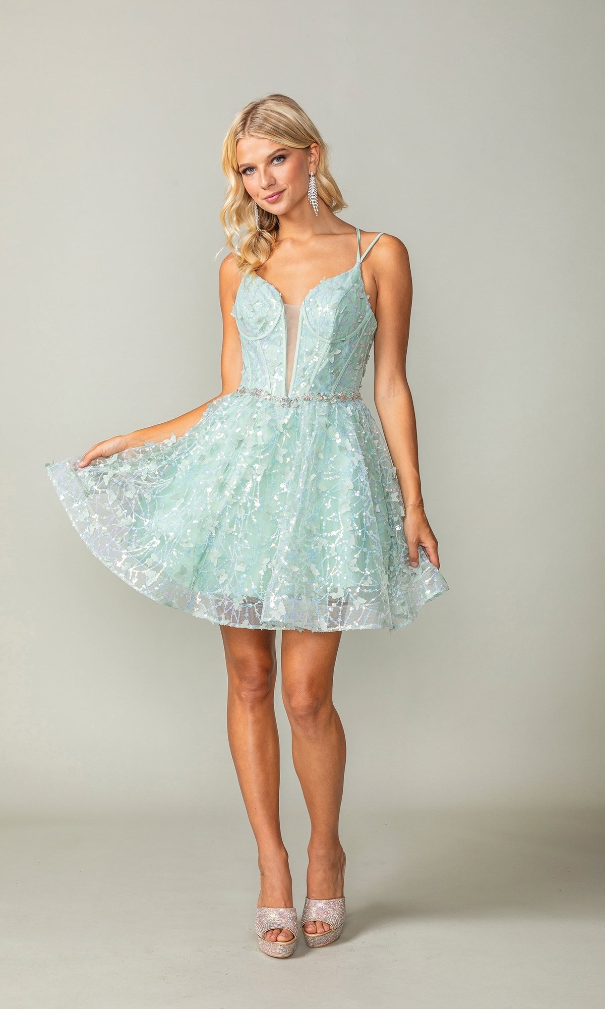 A woman poses against a plain backdrop in the Three-D Short Butterfly Homecoming Dress 3350—a light blue, sleeveless dress featuring a corset-style bodice and flared skirt—paired with high-heeled shoes.